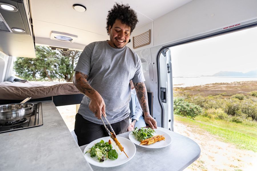 Een man is in de Mighty Highball camper aan het koken en serveert het eten op borden op de tafel