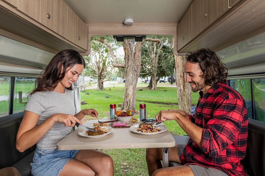 Een koppel is aan het eten in de zithoek achterin de Mighty Highball camper met de achterdeuren open met uitzicht op de natuur