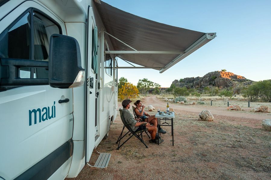 Een koppel zit op de campingstoelen onder de luifel van de Maui Sunset camper die geparkeerd staat in de indrukwekkende natuur van Australië