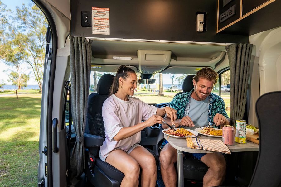 Een man en vrouw zitten op de draaibare bestuurdersstoelen en zijn aan het eten aan tafel in de Apollo Hitop camper in Australië