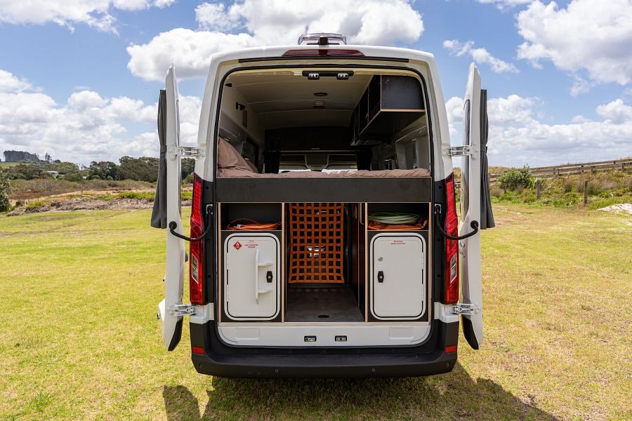 De Apollo Hitop camper staat geparkeerd op een grasveld in Nieuw-Zeeland met de achterdeuren open met bergruimte en daarboven het tweepersoonsbed