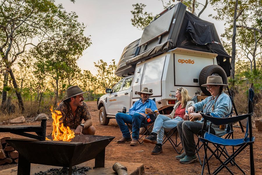 Een vriendengroep zit op de campingstoelen bij een kampvuur aan de achterkant van de Apollo 4WD Overland Camper in Australië