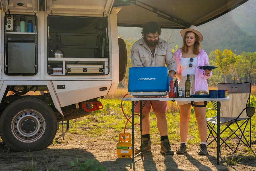 Een koppel is aan het koken in de buitenkeuken onder de luifel met de zijdeur van de Apollo 4WD Overland Camper op in de natuur van Australië