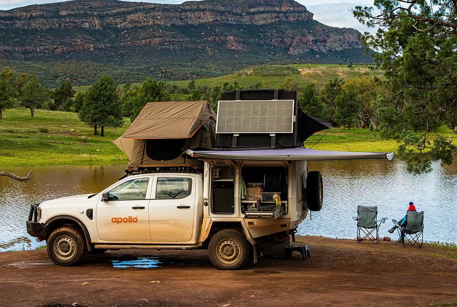 De Apollo 4WD Overland Camper staat met de daktenten en luifel open geparkeerd en een vrouw zit op de campingstoelen met uitzicht op een meer in Australië