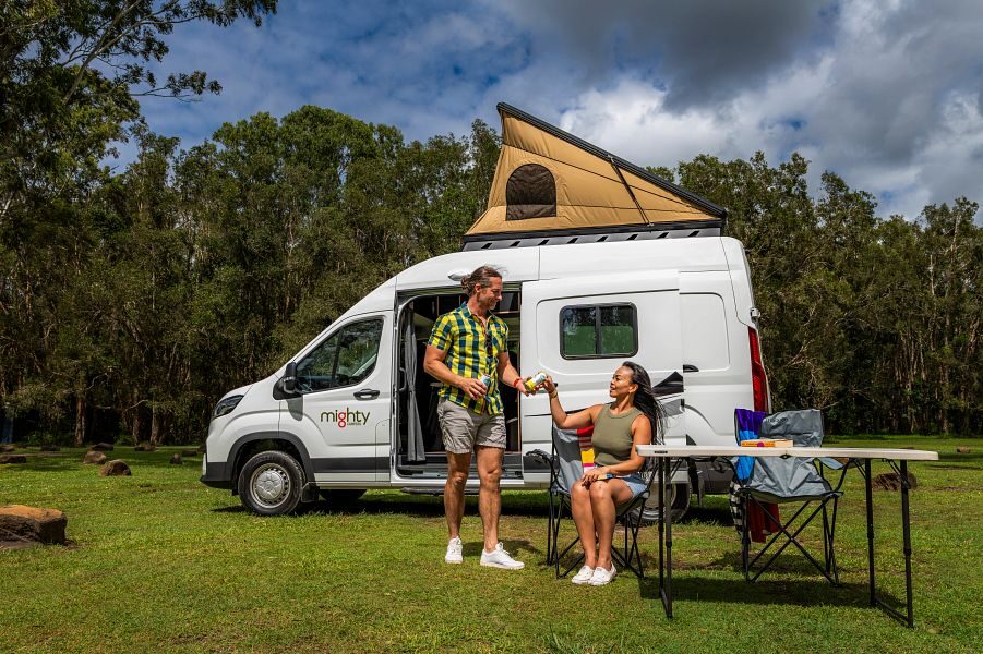 Een man geeft een blikje drinken aan zijn vrouw die op een campingstoel zit voor de Mighty Double Down camper die geparkeerd staat op het gras met de daktent open