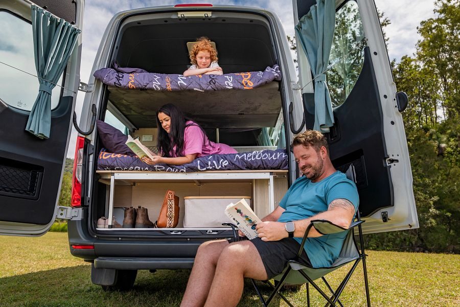 De achterkant van de Mighty Double Down camper in Australië waar een man in een campingstoel een boek zit te lezen, zijn vrouw op het onderste tweepersoonsbed ligt te lezen en een kindje in het bovenste bed ligt en naar buiten kijkt