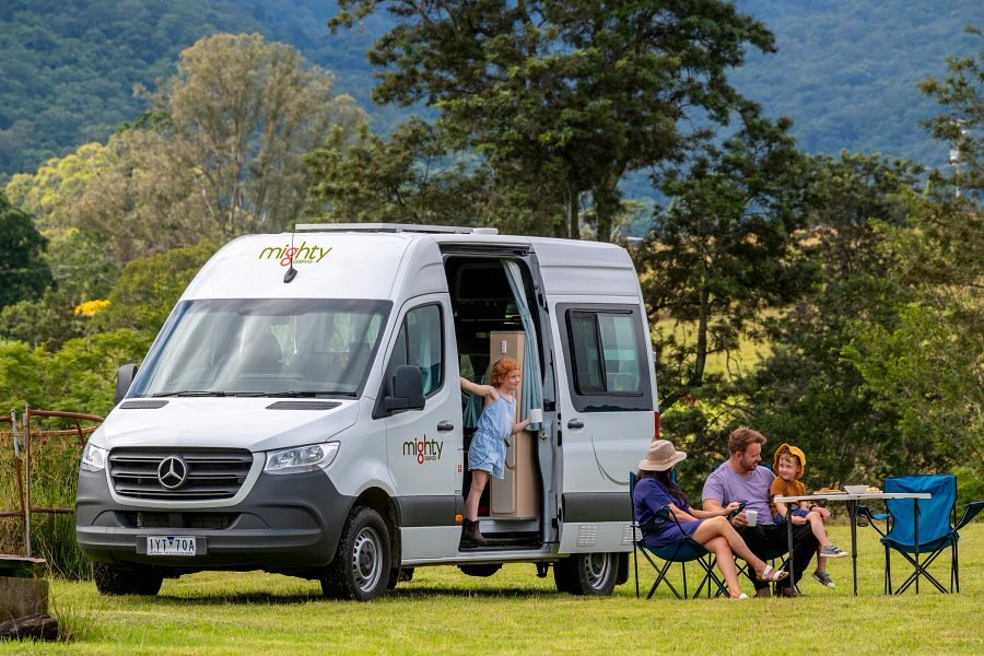 Een gezin zit op de campingstoelen voor de Mighty Double Down camper in de groene natuur van Australië terwijl een kindje in de deuropening van de camper staat