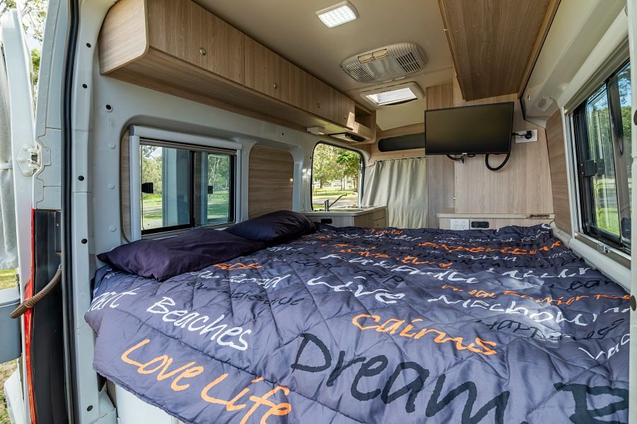 Een opgemaakt tweepersoonsbed achterin de Mighty Duo camper in Australië met kastruimte boven het bed en een TV