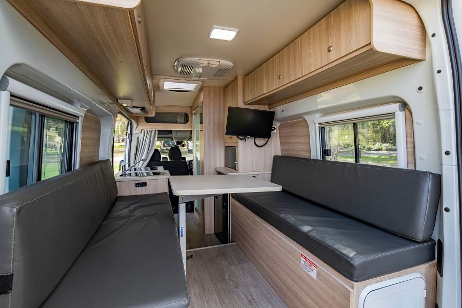 Vanuit de achterkant van de camper kijk je naar het interieur van de Mighty Duo met twee zitbanken, tafel, kastruimte en een TV