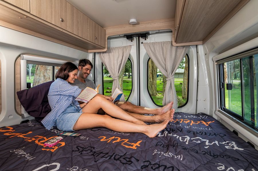 Een koppel zit op het tweepersoonsbed aan de achterkant van de Mighty Duo camper een boek te lezen