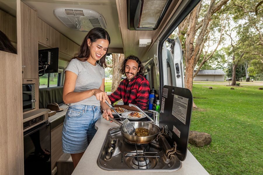 Een koppel is aan het koken in de keuken met de deuren open met uitzicht op de natuur in de Mighty Duo camper