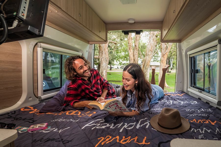 Een koppel ligt op het tweepersoonsbed en lachen om een boek die zij aan het lezen is met de achterdeuren open in de Mighty Duo camper