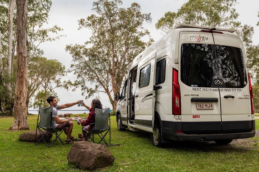 Een koppel zit naast de Mighty Duo camper op de campingstoelen en proosten samen met uitzicht op de natuur van Australië