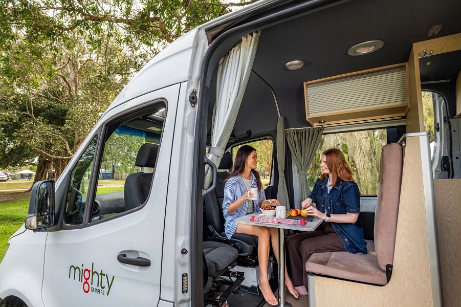Twee vriendinnen zitten aan tafel in de zithoek voorin de Mighty Deuce Plus camper met de zijdeur open