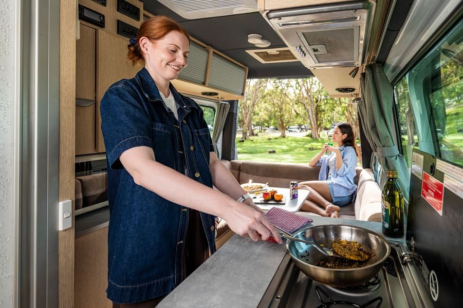 Een vrouw is aan het koken in de keuken van de Mighty Deuce Plus camper terwijl haar vriendin in de zithoek achterin de camper zit met de achterdeuren open