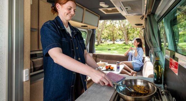 Een vrouw is aan het koken in de keuken van de Mighty Deuce Plus camper terwijl haar vriendin in de zithoek achterin de camper zit met de achterdeuren open