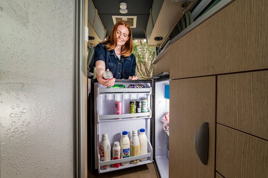 Een vrouw pakt een blikje drinken uit de openstaande koelkast in de keuken van de Mighty Deuce Plus camper