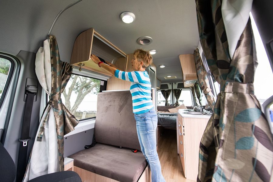 Een vrouw legt haar spullen in de opbergruimte boven de zitplaats in het midden van de Mighty Deuce Plus camper