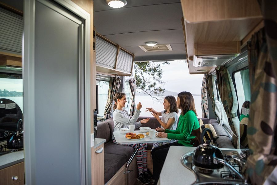 Een moeder zit met haar twee dochters te ontbijten aan tafel in de zithoek achterin de Mighty Deuce Plus camper met de achterdeuren op met uitzicht op het water