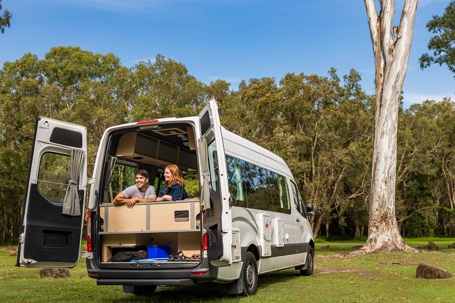 Een koppel kijkt naar buiten via de openstaande achterdeuren terwijl zij binnen in de Mighty Deuce Plus camper op bed zitten