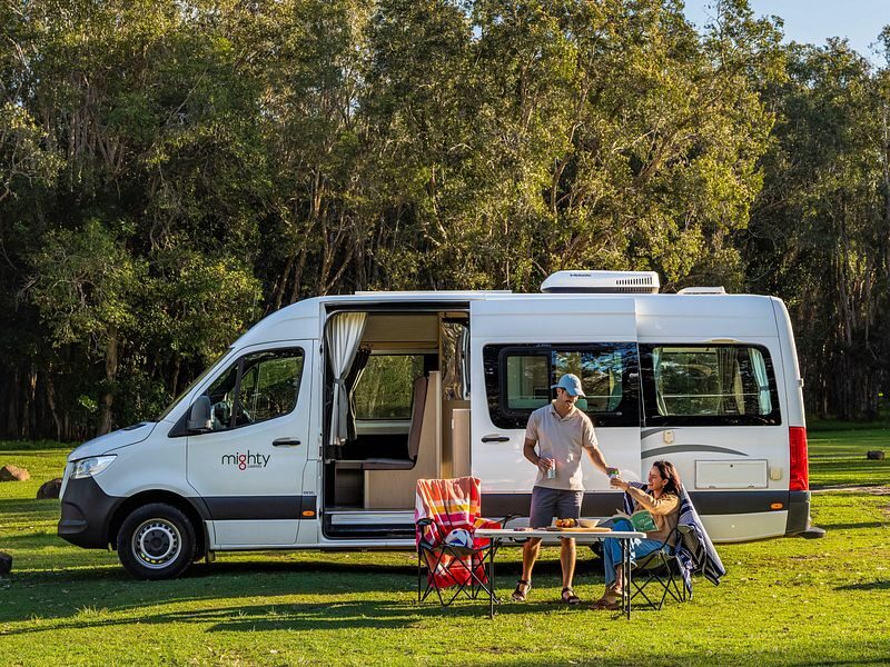 Een man geeft zijn vrouw wat te drinken terwijl zij op een campingstoel voor de Mighty Deuce Plus camper zit
