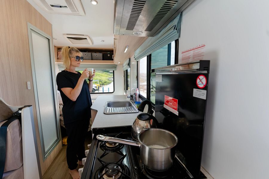 Een vrouw staat met haar drinken in de keuken van de Mighty Double Up camper terwijl er een pan met melk op de gaspit staat