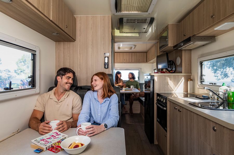 Een koppel zit met een mok met drinken en koekjes op tafel in het midden van de Mighty Double Up camper en achterin zit een ander koppel in de zithoek