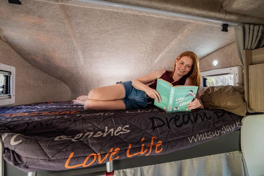 Een vrouw leest een boek in het tweepersoonsbed in de alkoof boven de cabine van de Mighty Double Up camper