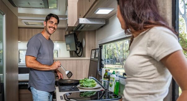 Een man is aan het koken en lacht naar zijn vrouw die in de deuropening van de Mighty Double Up camper staat