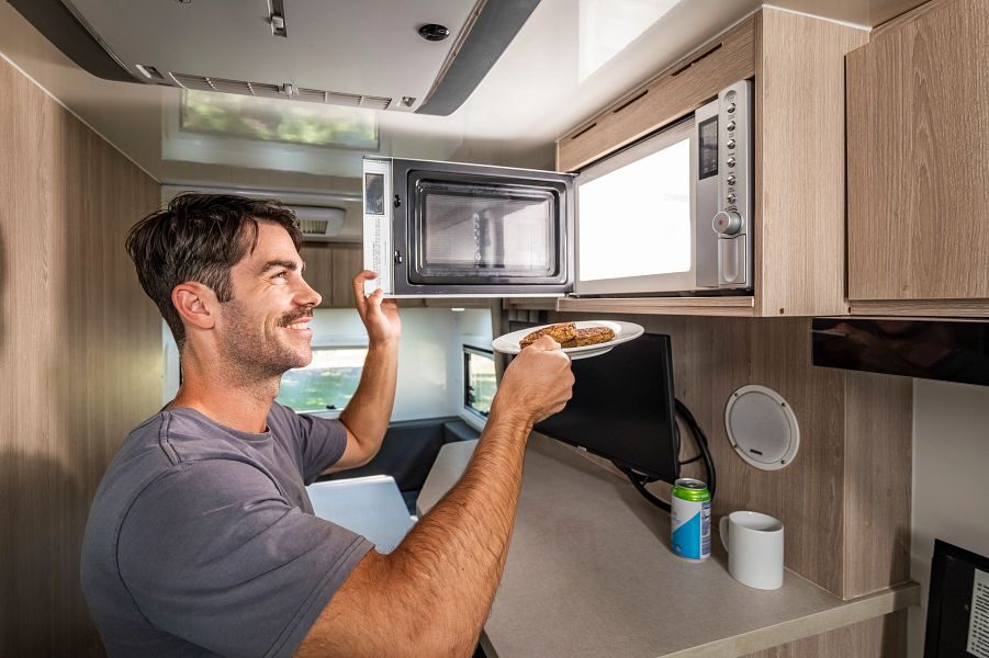 Een man doet de magnetron open en wil hier zijn bord met eten in doen in de keuken van de Mighty Double Up camper