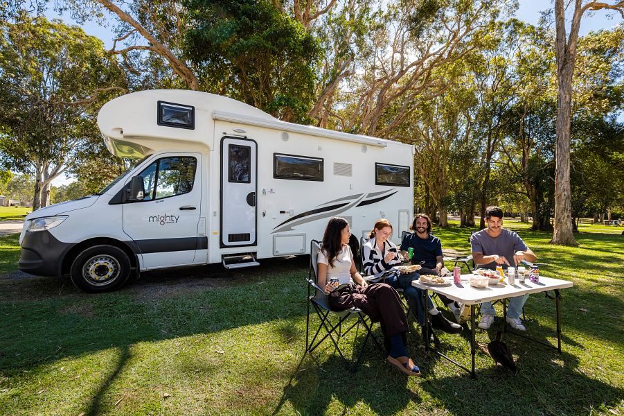 Een vriendengroep zit buiten op de campingstoelen aan tafel en achter hun staat de Mighty Double Up camper in het gras