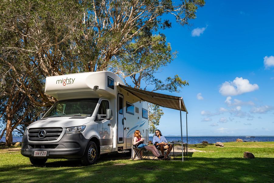 Een koppel zit aan tafel op de campingstoelen onder de luifel van de in het gras geparkeerde Mighty Double Up camper met uitzicht op het water
