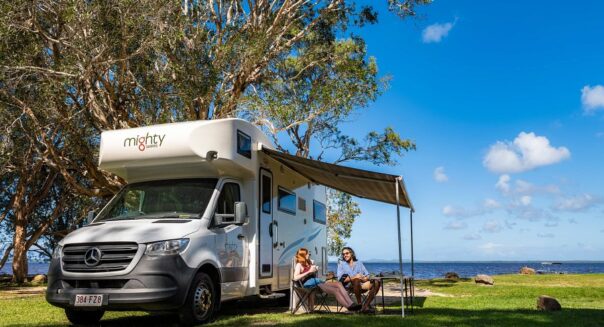 Een koppel zit aan tafel op de campingstoelen onder de luifel van de in het gras geparkeerde Mighty Double Up camper met uitzicht op het water