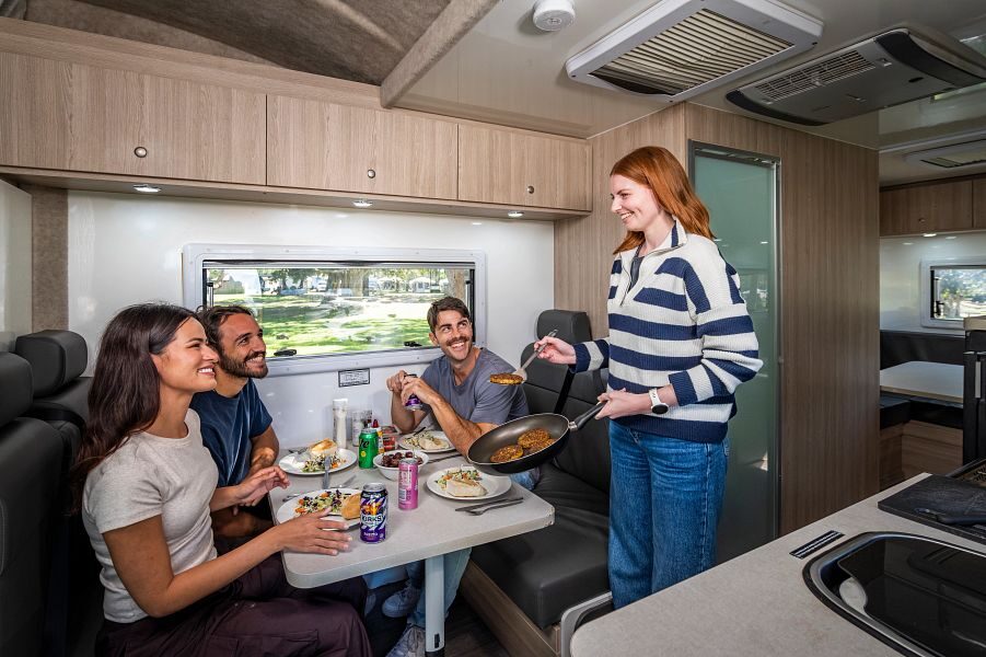 Een vrouw staat met een pan met gebakken hamburgers bij de tafel in het midden van de Mighty Big Six camper waar haar drie vrienden klaar zitten voor het diner