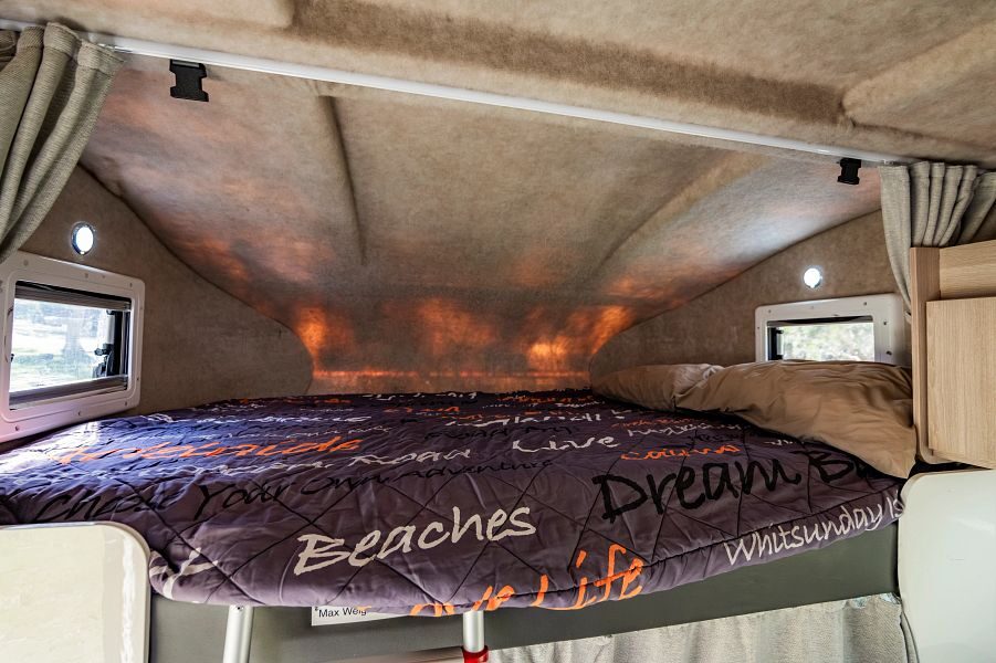 Het tweepersoonsbed in de alkoof met kleine ramen boven de cabine van de Mighty Big Six camper
