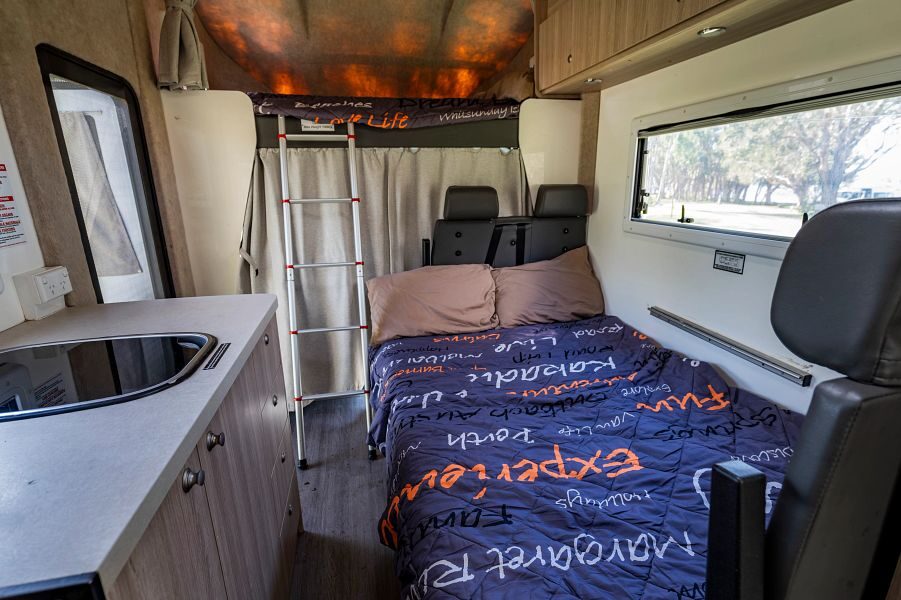 Een opgemaakt bed in het midden van de Mighty Big Six camper met daarachter een ladder naar het tweepersoonsbed in de alkoof