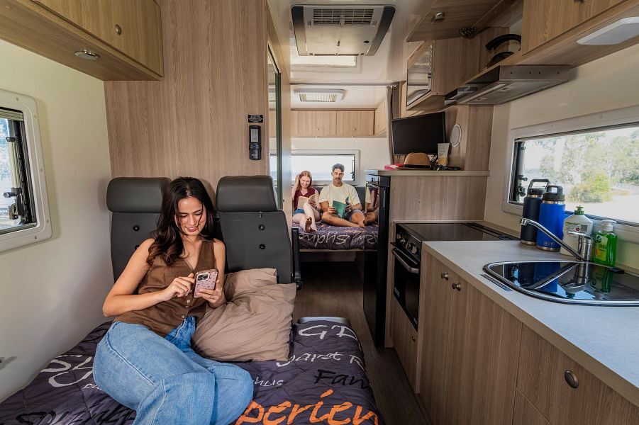 Een vrouw zit op haar telefoon en ligt op het bed in het midden van de Mighty Big Six camper met aan de achterkant een koppel die beide een boek aan het lezen zijn op het tweepersoonsbed