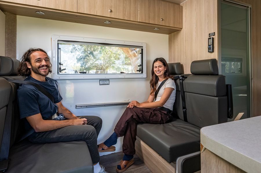 Een man en vrouw zitten tegenover elkaar met hun gordel om op de zitplaatsen achter de cabine tijdens het rijden in de Mighty Big Six camper