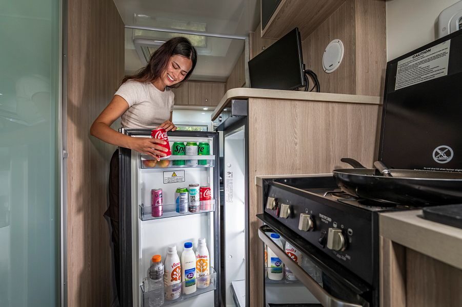 Een vrouw pakt een blikje drinken uit de koelkast in de keuken van de Mighty Big Six camper