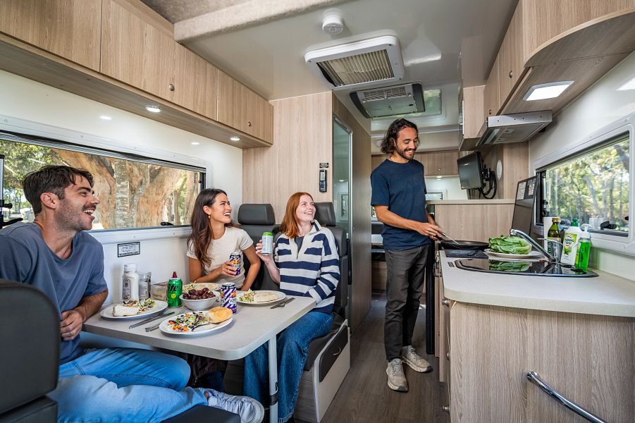 Een man staat in de keuken te koken terwijl drie vrienden zitten aan de tafel in het midden van de Mighty Big Six camper met een bord eten