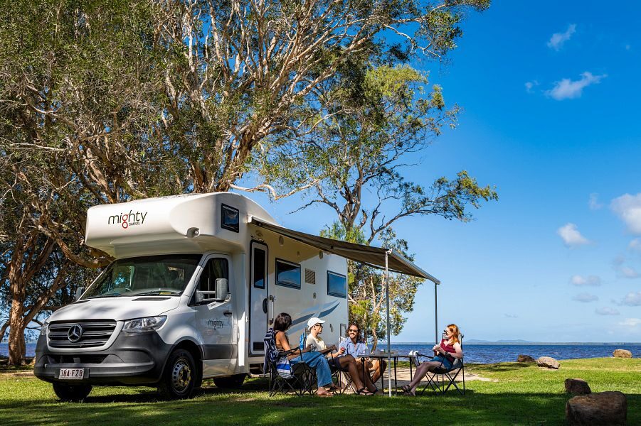 De Mighty Big Six camper staat geparkeerd op het gras bij de waterkant waar vier personen aan tafel zitten op de campingstoelen onder de luifel