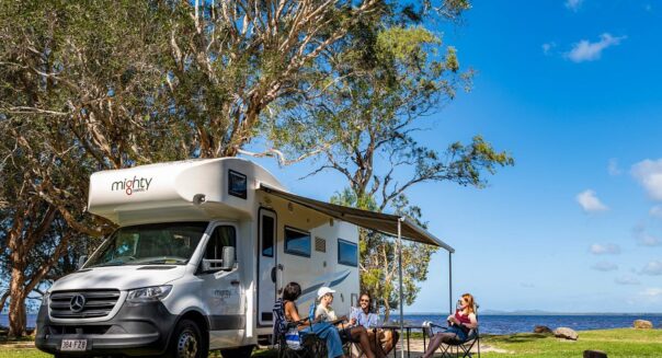 De Mighty Big Six camper staat geparkeerd op het gras bij de waterkant waar vier personen aan tafel zitten op de campingstoelen onder de luifel