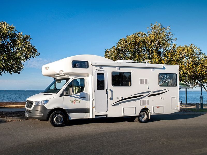 De zijkant van de Mighty Big Six camper die geparkeerd staat op een weg aan het water