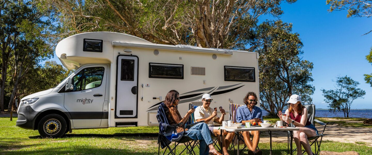 Twee stellen zitten buiten te kaarten met een Mighty camper op de achtergrond