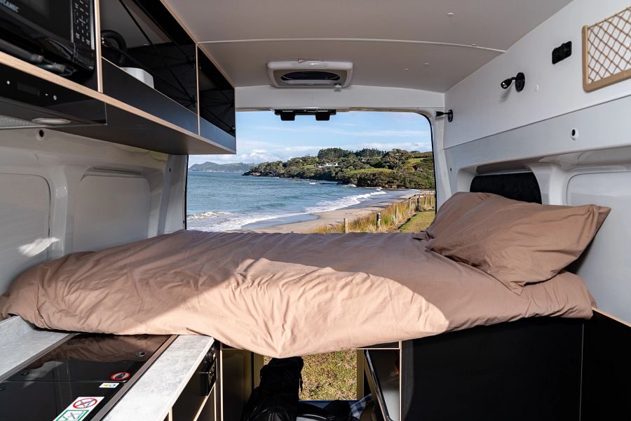 Een opgemaakt tweepersoonsbed achterin de Mighty Lite HiLITE camper met uitzicht op de kust van Nieuw-Zeeland