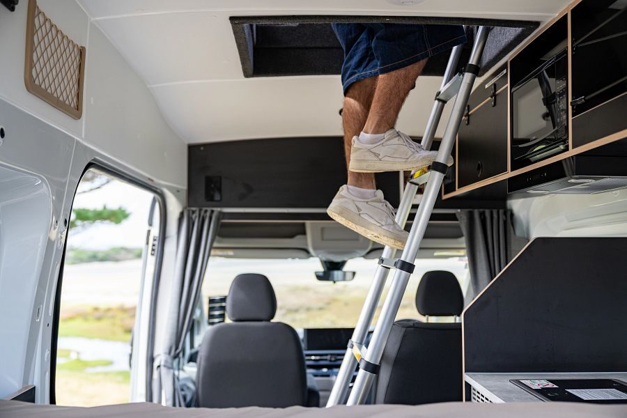 Een man staat op de ladder naar de daktent in de Mighty Lite DeLITE camper in Nieuw-Zeeland