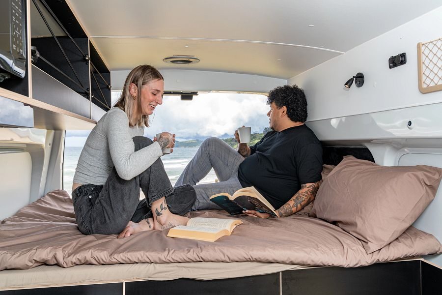 Een stel zit op het tweepersoonsbed achterin de Mighty Lite DeLITE camper wat te drinken met de achterdeur open met uitzicht op de zee in Nieuw-Zeeland