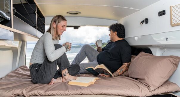 Een stel zit op het tweepersoonsbed achterin de Mighty Lite DeLITE camper wat te drinken met de achterdeur open met uitzicht op de zee in Nieuw-Zeeland