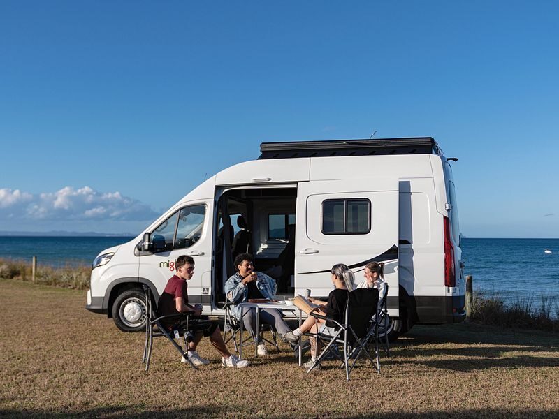 Een vriendengroep zit voor de Mighty Lite DeLITE camper met uitzicht op de zee in Nieuw-Zeeland