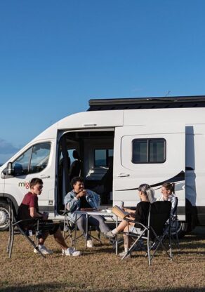 Een vriendengroep zit voor de Mighty Lite DeLITE camper met uitzicht op de zee in Nieuw-Zeeland